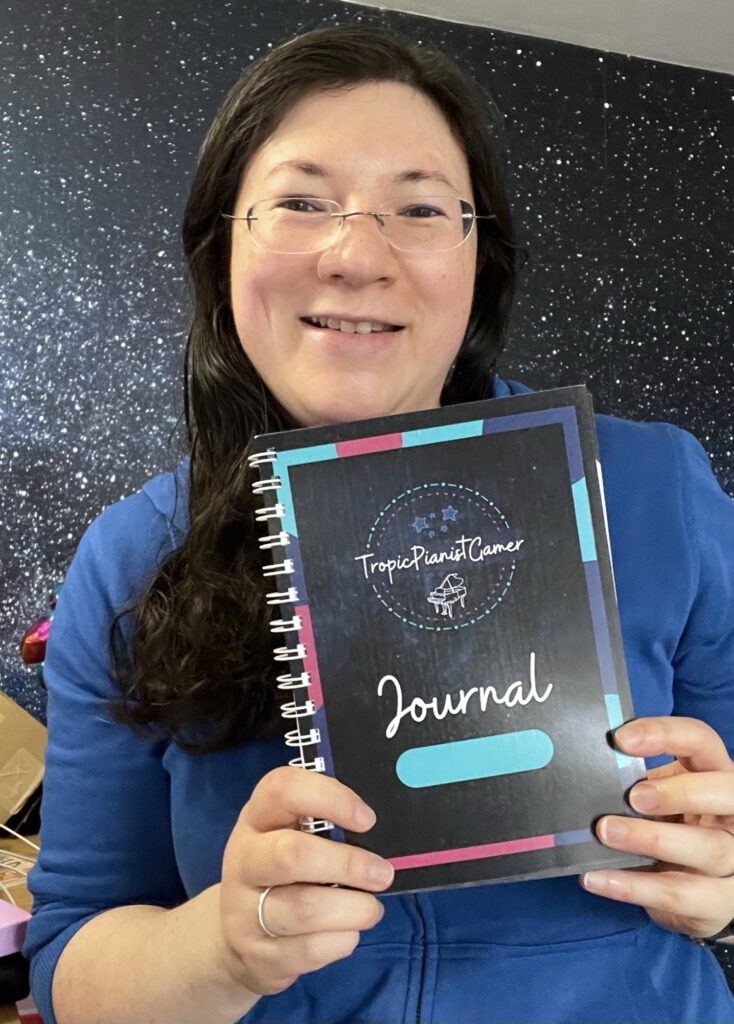 Jen is pictured in a royal blue hoodie, holding a spiral bound version of her self published journal.