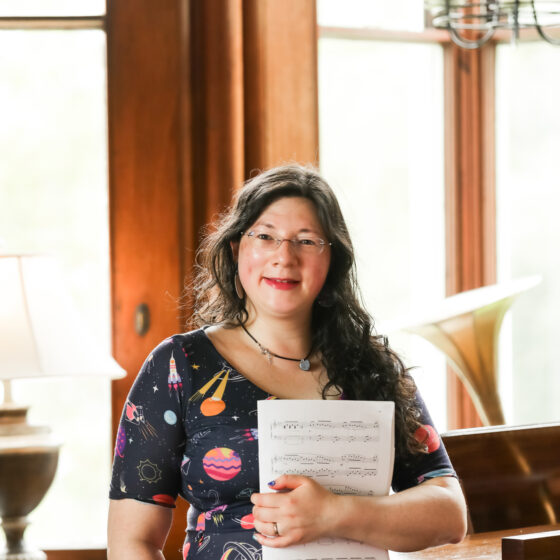 A picture of Jen stood by Chateau Rhianfa's Grand piano, holding a book of music. Wearing a space themed Popsy Dress, with long wavy hair.