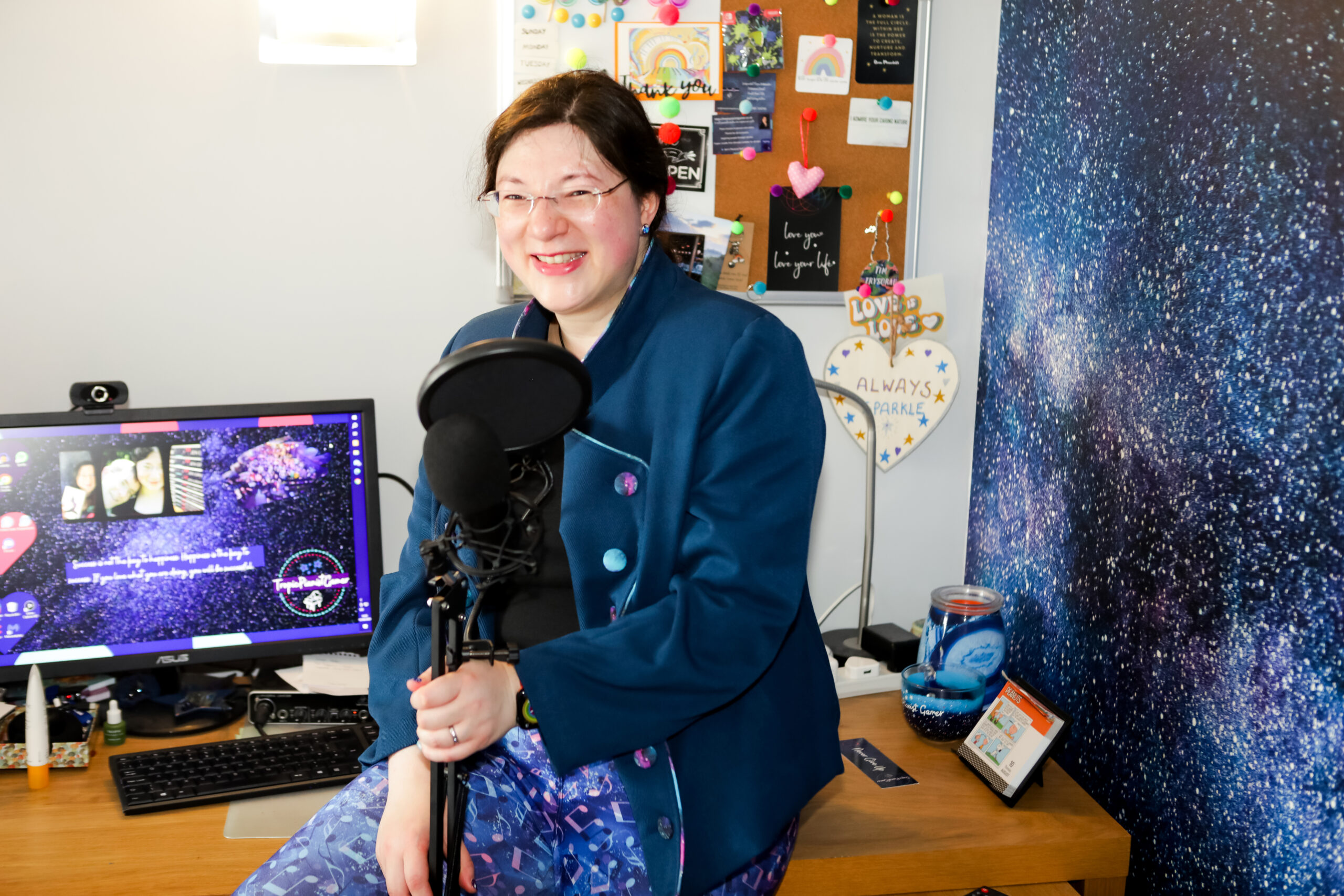 Jen is perched on her desk laughing at the floating microphone she's holding. Computer with desktop wallpaper on in the background, with feature galaxy themed wallpaper on the right.