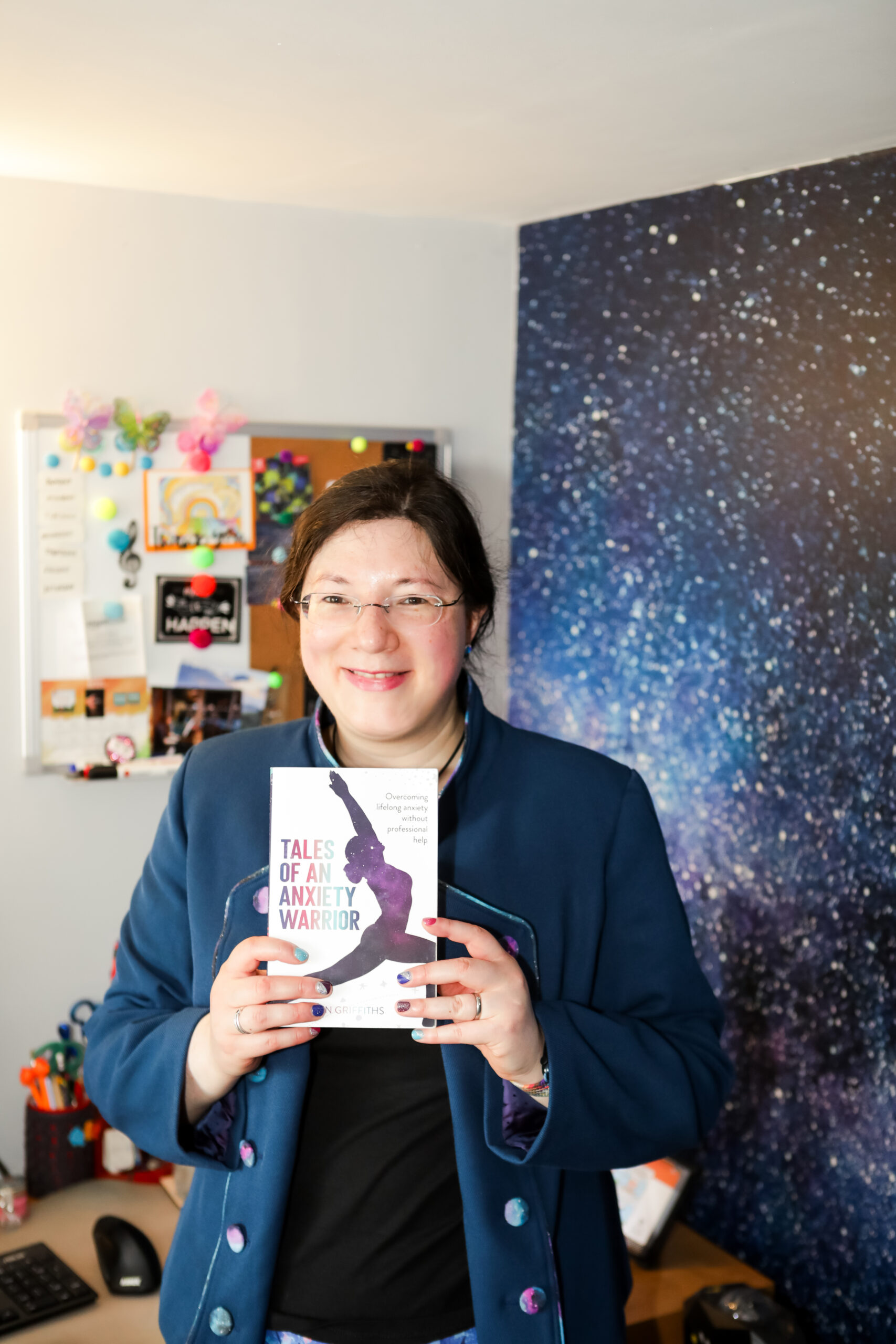 My Books 1 Jen is smiling while holding her book up. She is by her galaxy themed wallpaper, with her work station in the backround. She is wearing her custom made galaxy themed jacket.