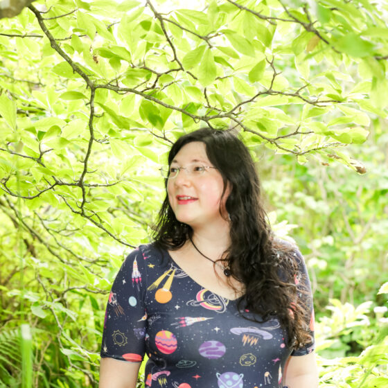 Jen is in her space dress, gazing into the distance in a very green leafy area.