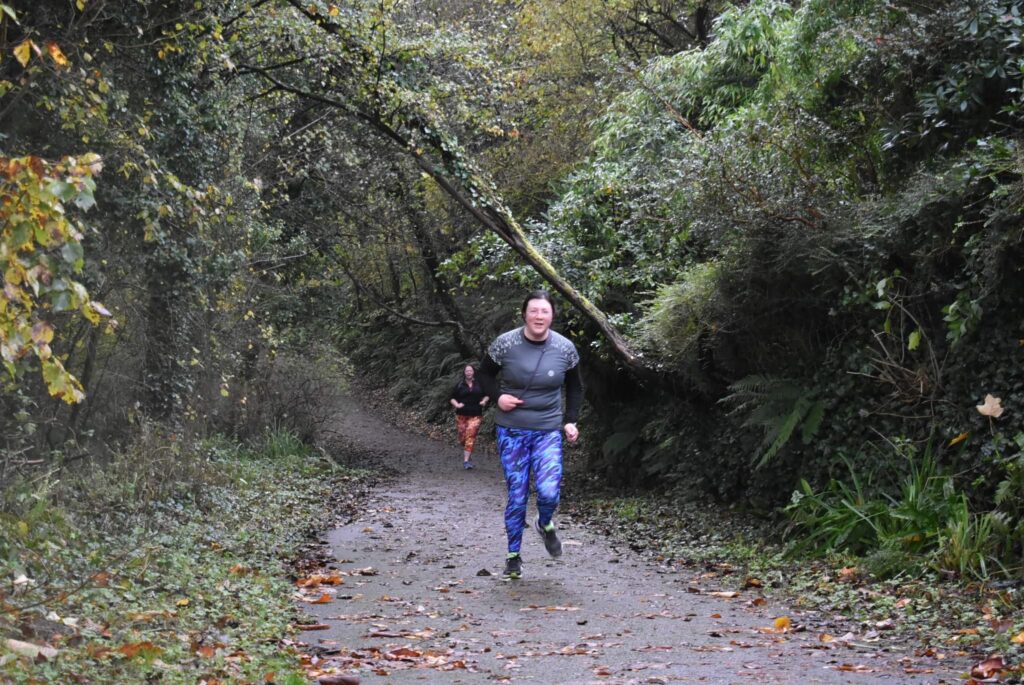 Nant y Pandy Parkrun 11/11/23 2 Jen doing parkrun at Nant y Pandy. Wearing blue lava active LLL leggings, and a grey reflective running top. Lady in LLL orange leggings in background. Woodland scenery.