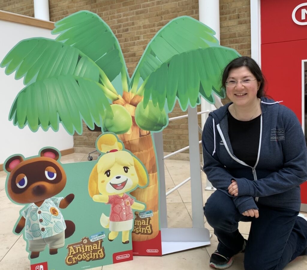 Me crouched next to Isabelle and Tom Nook by a palm tree (cardboard standee) smiling for the camera in my navy blue NNW hoodie. backdrop is the official Nintendo HQ door.