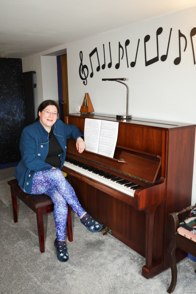 Jen's Musical Self Care Den - A Fun and inspiring place to be! 1 Jen sat at her piano, ready to entertain Jen's Musical Self Care Den with her piano music. Wearing her galaxy themed jacket and musical leggings.