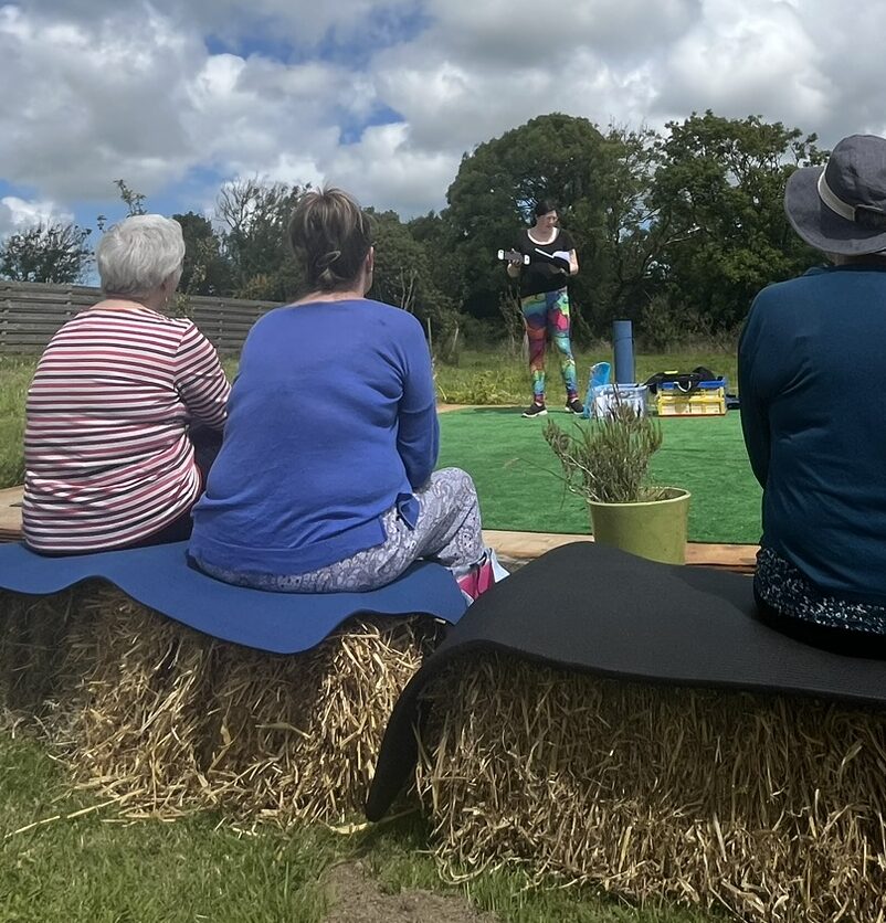 Speaker at a Brand New Yoga Festival- June 2024 - a Major 2024 Goal accomplished! 1 Jen giving a talk in the background while the group watched.