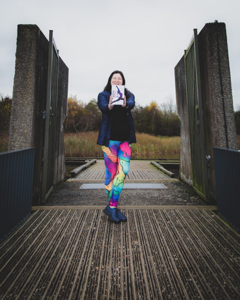a posed shot of me in my rainbow leggings with navy sparkly jacket, smiling and holding out my book. Stood at the end of the bridge. 