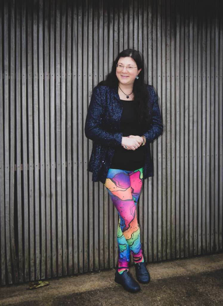 A posed shot of me in rainbow leggings, navy sparkly jacket smiling and glancing sideways, against a wooden structure. 