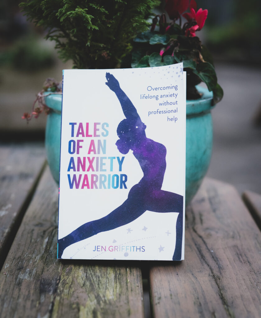 My book "Tales of an Anxiety Warrior" leaning against a green plant pot with red flowers in it. 