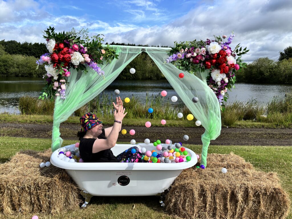 Festivals of 2025 Fulfilled & 3 Public Speaking Goals Smashed! 2 picture of me in a bathtub with plastic balls, throwing the balls into the air. Wearing rainbow feather bucket hat.